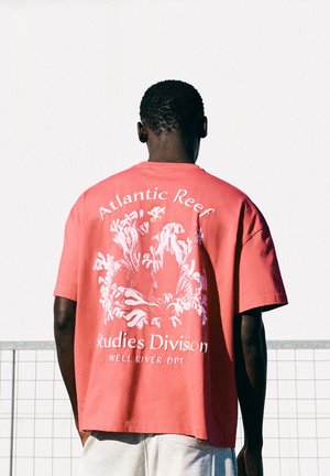Mann, der ein korallenrotes T-Shirt mit weißem Korallenriff-Design und dem Text "Atlantic Reef Studies Division Well River DPT" auf dem Rücken trägt, steht draußen.