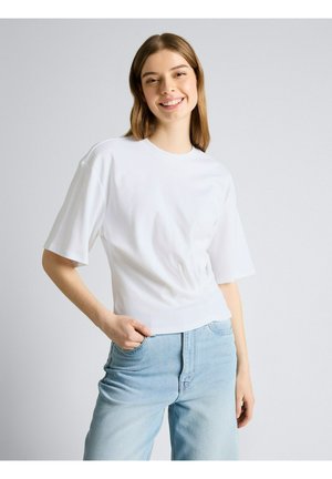 Młoda kobieta uśmiechająca się, ubrana w prostą białą koszulkę z krótkim rękawem i jasnoniebieskie, wysokie dżinsy, z ręką w kieszeni na jednolitym tle.