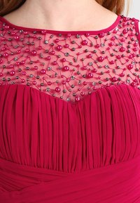 Fuchsia klänning med en plisserad överdel, prydd med en genomskinlig overlay av pärlor med rosa och metalliska pärlor, samt en rundad halsringning.