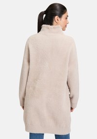 Beżowy, puszysty kardigan z wysokim kołnierzem, długimi rękawami i luźnym krojem, charakteryzujący się miękką fakturą i wydłużonym tyłem.