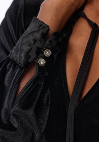 Czarna aksamitna odzież z wzorzystym kołnierzem. Posiada dwa dekoracyjne guziki z srebrnymi akcentami oraz głęboki dekolt w kształcie V.