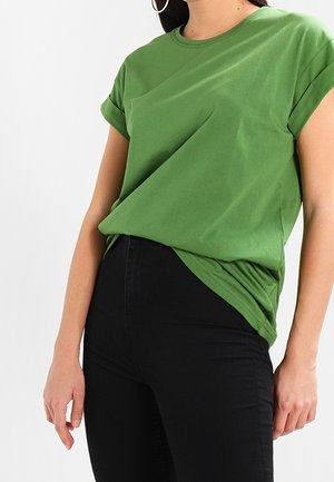 Femme portant un t-shirt vert à manches courtes légèrement rentré dans un pantalon noir taille haute, debout devant un fond blanc.