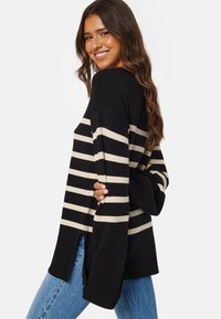 Maglione a righe nero e crema con vestibilità ampia, maniche larghe e spacchi laterali, abbinato a jeans in denim blu chiaro.