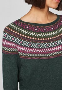 Maglione verde scuro con un colletto decorato da motivi geometrici in rosa, bianco e arancione. Texture morbida con scollatura rotonda.