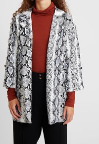 Slangenprintjas in zwart en wit met blauwe accenten; losse pasvorm, open voorkant en brede mouwen. Gelaagd over een roestkleurige coltrui.