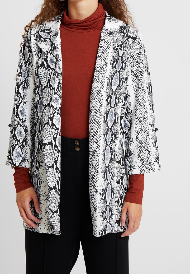 Slangenprintjas in zwart en wit met blauwe accenten; losse pasvorm, open voorkant en brede mouwen. Gelaagd over een roestkleurige coltrui.