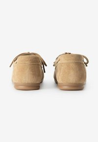 Chaussons en daim beige clair avec une texture douce. Présentent une semelle basse et flexible ainsi que des lacets décoratifs à l'arrière pour un ajustement.