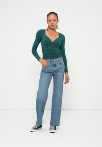 Grünes Langarmoberteil mit V-Ausschnitt und gerafften Seiten, kombiniert mit hellblauen Jeans im geraden Schnitt und schwarzen Sneakers. Einfarbiger weißer Hintergrund.