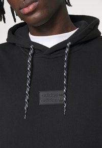 Black hoodie with a textured fabric, featuring a logo patch, and black-and-white drawstrings with a braided pattern.