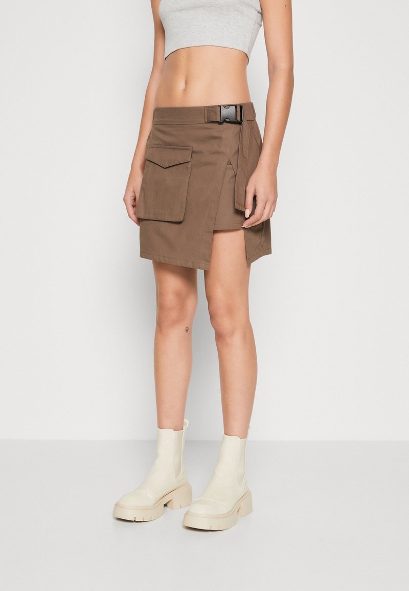 Femme portant une jupe portefeuille marron avec une poche latérale et une boucle noire, associée à un crop top gris et des bottines blanches.