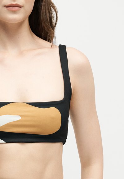Γυναίκα που φοράει μαύρο crop top με φαρδιές τιράντες και αφηρημένο καφέ και λευκό μοτίβο μπροστά, εμφανίζεται από τους ώμους ως τη μέση.
