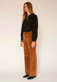 Blouse noire à manches longues avec des coutures décoratives ; pantalon large en velours côtelé marron ; bottines noires ; bracelets empilés.