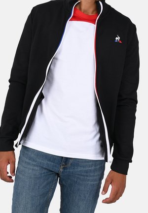 Sort lynlåstrøje med hvide detaljer og en rød og blå kant, båret over en rød og hvid skjorte, kombineret med blå jeans.