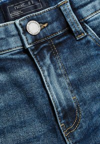 Vaqueros de mezclilla en azul oscuro con costuras amarillas, que presentan un botón plateado con marca y una etiqueta con "next . 82" en la cinturilla.