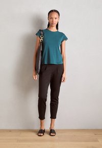T-Shirt in Türkis mit kurzen Ärmeln und Spitzenbesatz, schwarze schmale Hose und schwarze Rutschsandalen. Das Modell trägt eine große schwarze Umhängetasche.