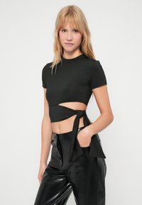 Top scurt negru cu maneci scurte, prezentând un detaliu legat pe lateral și o decupare, asortat cu pantaloni negri lucioși, cu talie înaltă.