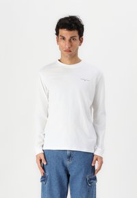 T-shirt blanc à manches longues avec un col rond et un petit logo "TOMMY JEANS" sur la poitrine, associé à un pantalon cargo bleu avec des poches latérales.