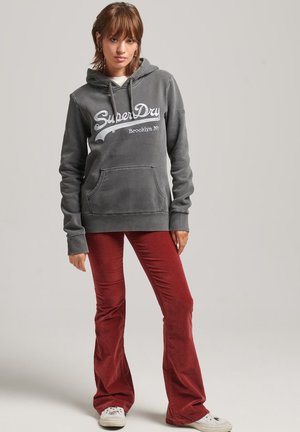 Felpa grigia in cotone con tasca frontale e cappuccio con cordino; presenta il logo bianco "SuperDry". Abbinata a pantaloni rossi a campana e sneaker bianche.