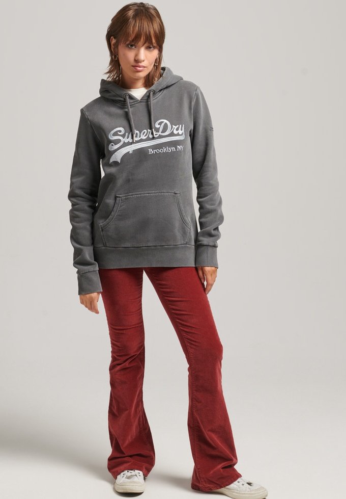 Hanorac gri din bumbac cu un buzunar frontal și glugă cu șnur; are logo-ul alb "SuperDry". Asortat cu pantaloni evazați roșii și adidași albi.