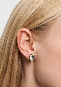 Nærbillede af et venstre øre med lige blond hår, iført en lille hjerteformet ørering med en gylden kant og reflekterende sølvcentrum.