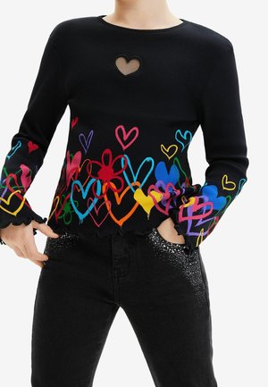 Zwart geribbeld top met lange mouwen, voorzien van een hartvormige uitsparing en kleurrijke hartpatronen langs de zoom en mouwen. Gecombineerd met versierde zwarte jeans.