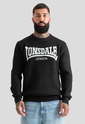 Homme avec barbe et tatouages au cou portant un pull noir Lonsdale London et un jean bleu clair, debout devant un fond uni.