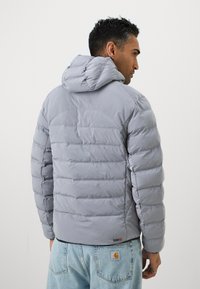 Grå pufferjakke med hætte, med vandrette quiltede sektioner og elastiske manchetter, båret over lyseblå denimjeans med et logo-patch.