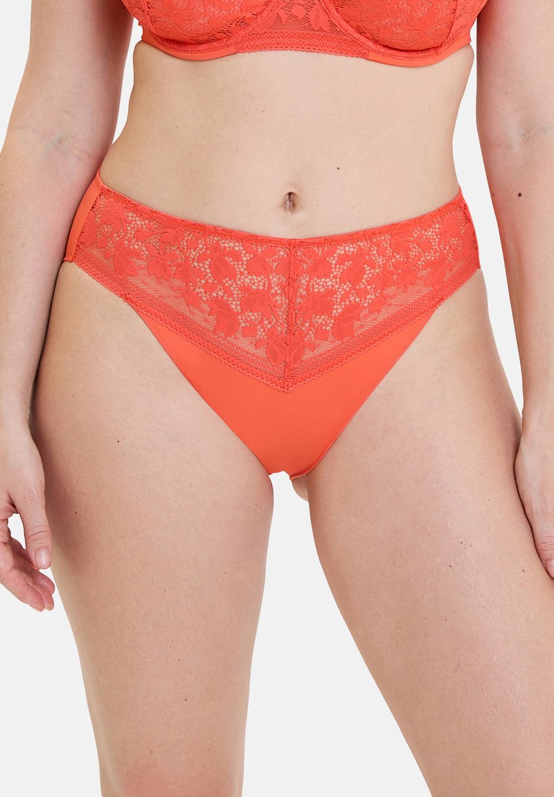 Donna che indossa slip bikini in pizzo corallo a vita alta e reggiseno abbinato, in piedi con le braccia rilassate contro uno sfondo bianco.