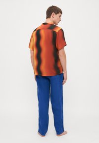 Camisa de manga corta en un patrón degradado de naranja y negro, combinada con pantalones de lino azules. La camisa presenta un collar y un corte relajado.