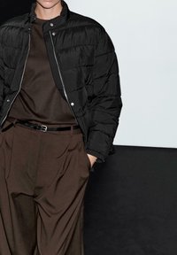 Person wearing a black quilted jacket over a brown shirt and high-waisted brown pleated pants with a black belt, with one hand in pocket.