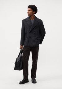 Dubbelzijdige donkergrijze getailleerde overjas, geweven stof, gecombineerd met donkerbruine corduroy broek en zwarte leren schoenen, met een zwarte tas in de hand.
