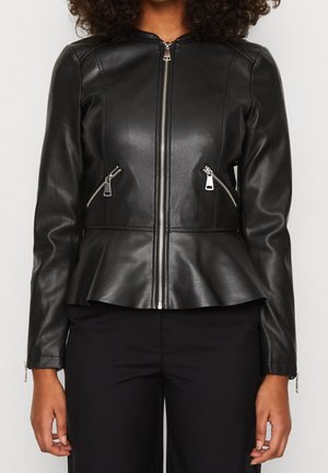 Vrouw draagt een zwart leren jack met perforaties, een zilveren ritssluiting aan de voorkant, ritssluitingen op de zijkanten en een uitlopende zoom, gecombineerd met zwarte broek.