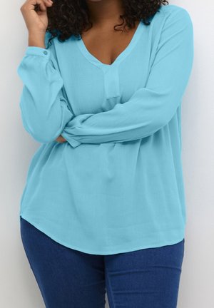 Kvinde med krøllet hår iført en lyseblå langærmet V-hals bluse og mørkeblå bukser, med armene krydsede, stående mod en hvid baggrund.