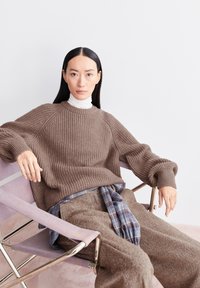 Bruine geribde trui met raglanmouwen, laagjes over een witte turtleneck. Losse bruine broek en een blauw geruite sjaal maken de outfit compleet.
