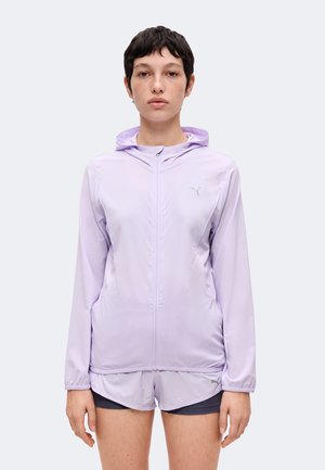 Jeune femme aux cheveux courts et foncés portant une veste à capuche violet clair zippée et un short assorti, debout devant un fond uni.