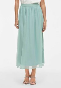 Světle tyrkysová maxi sukně vyrobená z lehkého chiffonového materiálu, se sbíraným pasem a měkkou, průsvitnou texturou. V kombinaci se stříbrnými sandály na tenkých páscích.