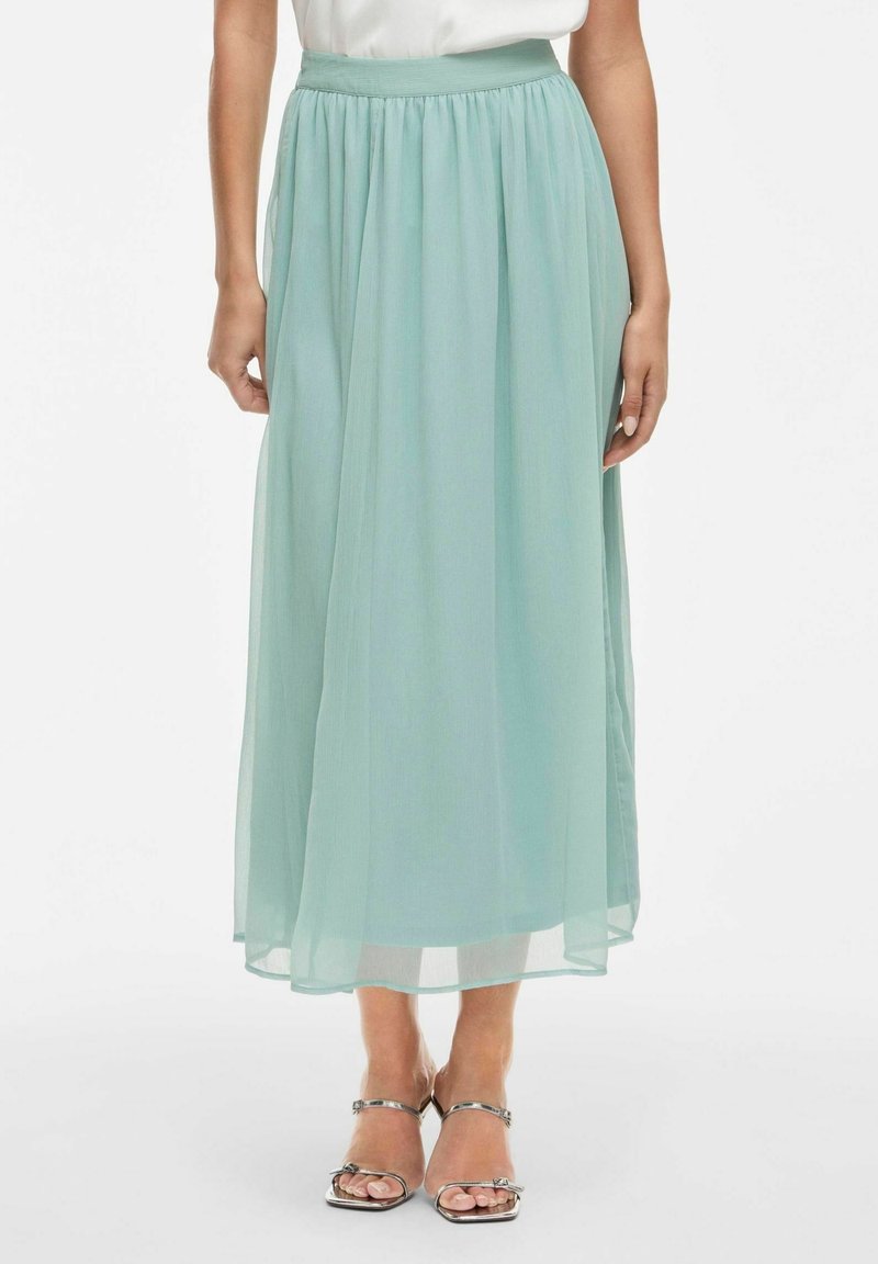 Světle tyrkysová maxi sukně vyrobená z lehkého chiffonového materiálu, se sbíraným pasem a měkkou, průsvitnou texturou. V kombinaci se stříbrnými sandály na tenkých páscích.