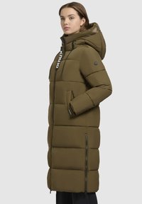 Olivovo zelený dlhý puffový kabát s kapucňou, so zipsom na prednej strane, detailom loga a bočnými vreckami. Ukončený prešívaným dizajnom.