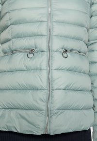 Doudoune vert clair avec un matelassage horizontal, des fermetures éclair argentées, et deux poches zippées à l'avant. Texture lisse et légèrement brillante.