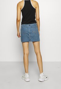 Kvinna som står vänd bort ifrån kameran, iklädd svart linne, blå denim-minikjol med råa kanter, vita sneakers och sockar mot en enfärgad bakgrund.