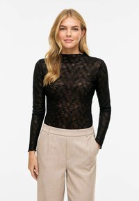 Czarna bluzka z koronki z długim rękawem i wysokim kołnierzem, ozdobiona misternymi kwiatowymi wzorami. Stylizowana z beżowymi, dopasowanymi spodniami.