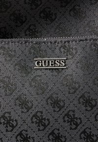 Črna tkanina denarnice z izbočeno srebrno logotipom "GUESS" in teksturiranim vzorcem fleur-de-lis po celotni površini.