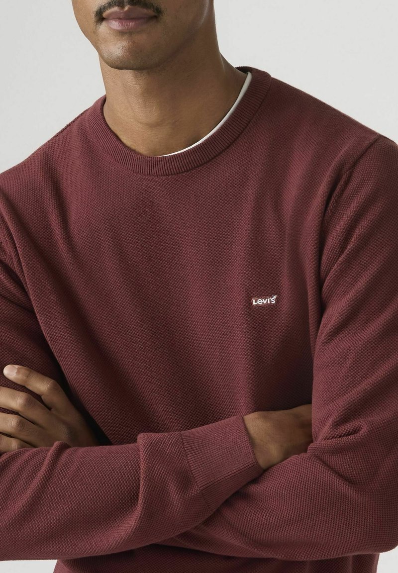 Hombre con los brazos cruzados, vistiendo un suéter de manga larga texturizado color granate de Levi's sobre una camisa blanca de cuello redondo, frente a un fondo liso.