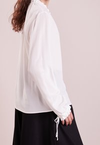 White long-sleeve blouse with gathered neckline and cuffs, featuring a lightweight, smooth fabric; paired with a black skirt.