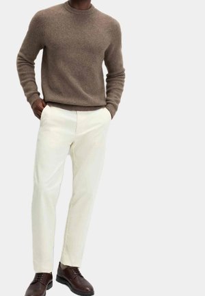 Homme portant un pull en laine marron, un pantalon écru et des chaussures en cuir marron foncé, debout les mains dans les poches.