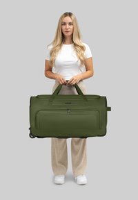 Grüne Reisetasche mit zwei oberen Griffen, Reißverschlüssen und Fronttasche, aus strapazierfähigem Stoff, präsentiert von einer Person in neutraler Kleidung.