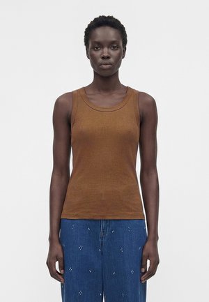 Femme aux cheveux courts et bouclés portant un débardeur marron sans manches et un jean bleu avec de petits motifs blancs, debout devant un fond blanc uni.