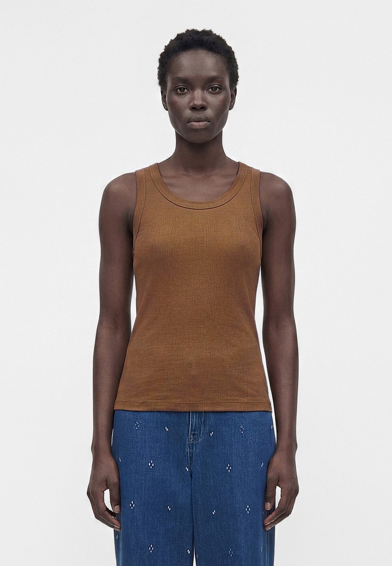 Γυναίκα με κοντά σγουρά μαλλιά που φοράει ένα καφέ αμάνικο τοπ και μπλε τζιν με μικρά λευκά σχέδια, στέκεται μπροστά σε ένα απλό λευκό φόντο.