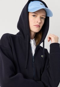 Sudadera con cremallera en color azul marino, con una tela texturizada, cordones ajustables y un logotipo verde en el frente. Combinada con una gorra de color azul claro.