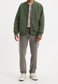 Hombre con chaqueta bomber verde, camisa a rayas beige y azul, jeans grises y zapatillas blancas de pie contra un fondo liso.
